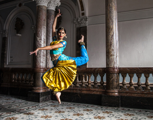 South Asian dancer wearing traditional blue and gold trousers and cropped top with bells on the ankles and a gold jewellery headpiece. Jumping in the air with point toe and one arm above the head and the other crossed over to the left. In front of the museum marble staircase. South Asian dancer wearing traditional blue and gold trousers and cropped top with bells on the ankles and a gold jewellery headpiece. Jumping in the air with point toe and one arm above the head and the other crossed over to the left. In front of the museum marble staircase.