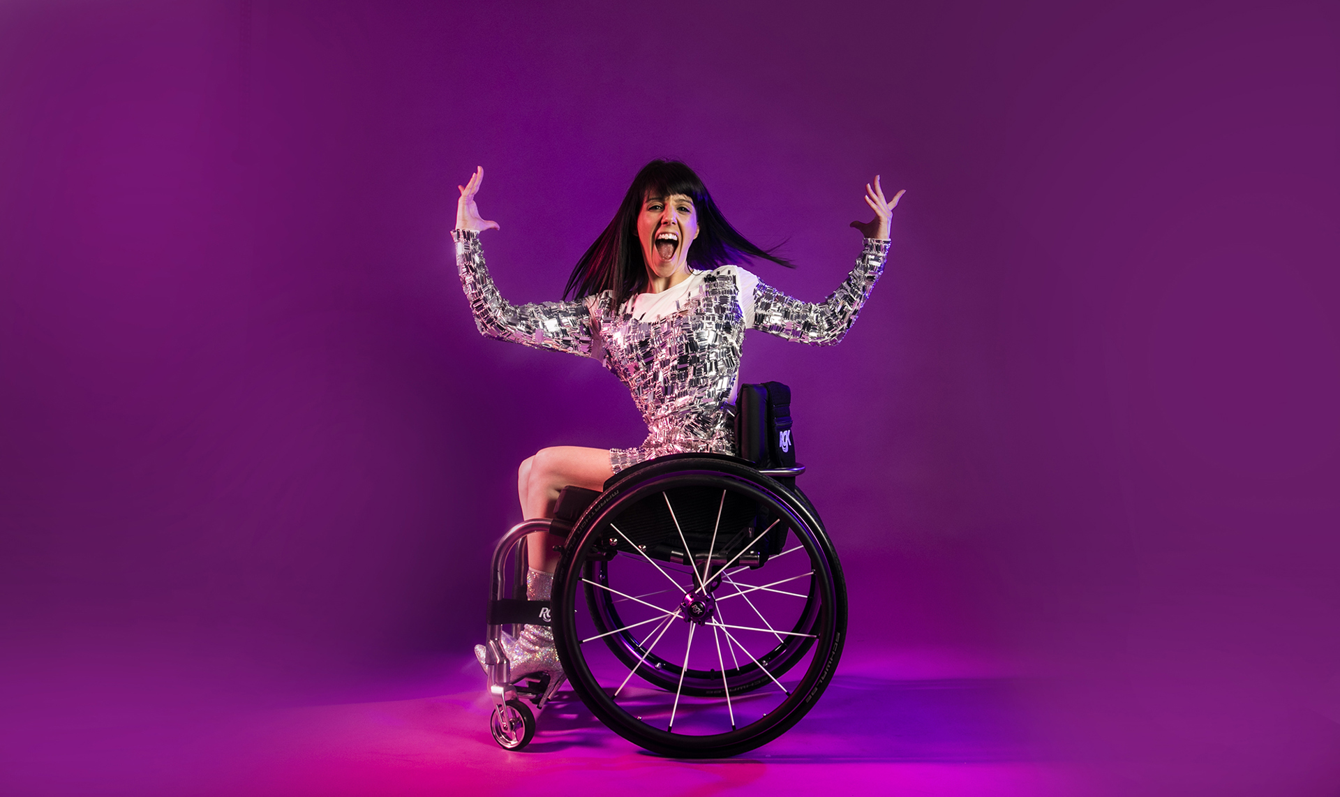 White female wheelchair dancer with black hair. With arms above her head and wheel chair side on. Facial expression wide mouth excited. Wearing silver sparkle dress and silver high heeled shoes in front of hot pink studio background.