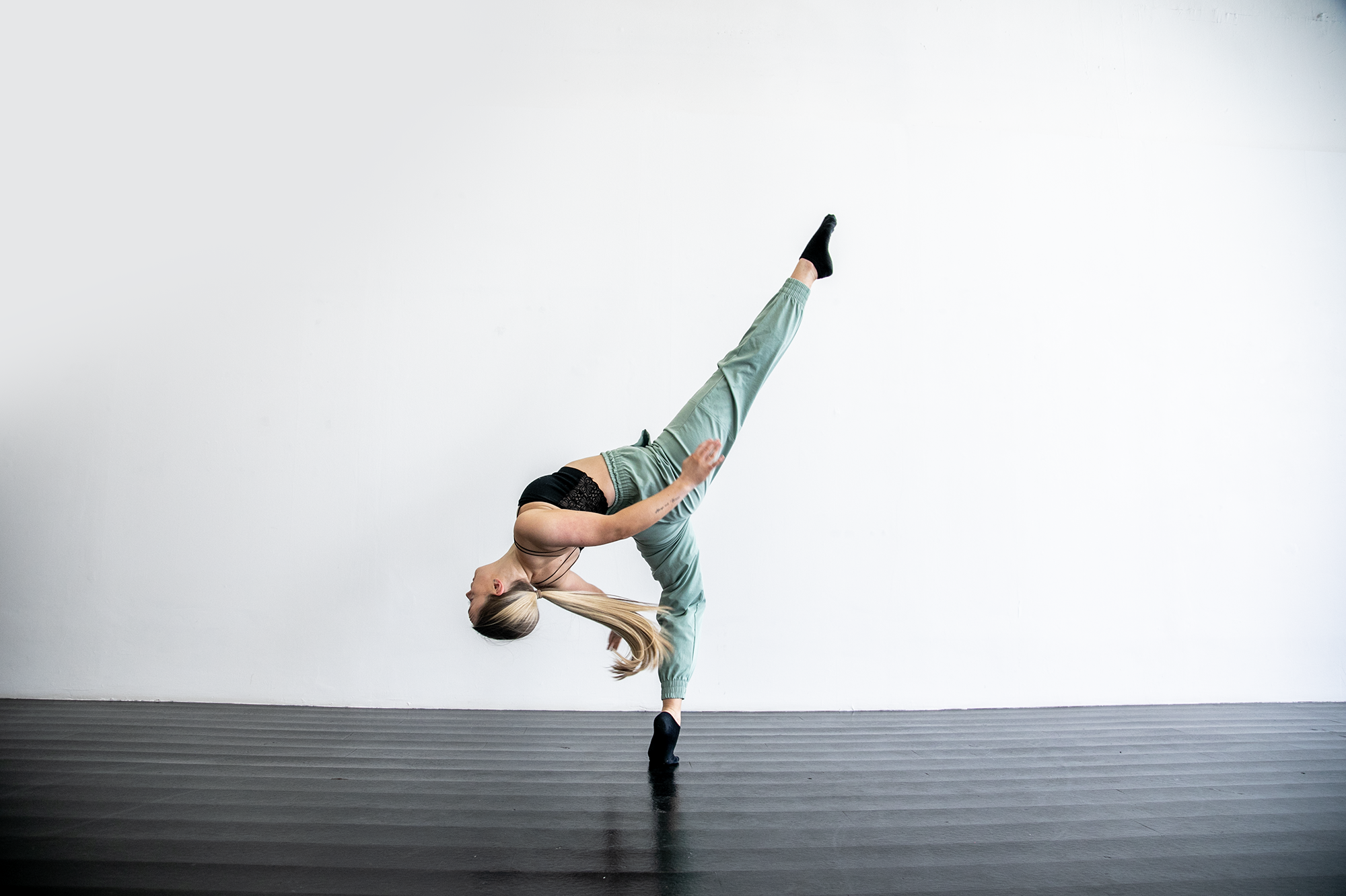 blonde white female in dance studio with black floors. One leg kicked high, other on demi point, leaning back with arms behind. Wearing blak strap top and green trousers and long blonde ponytail blonde white female in dance studio with black floors. One leg kicked high, other on demi point, leaning back with arms behind. Wearing blak strap top and green trousers and long blonde ponytail