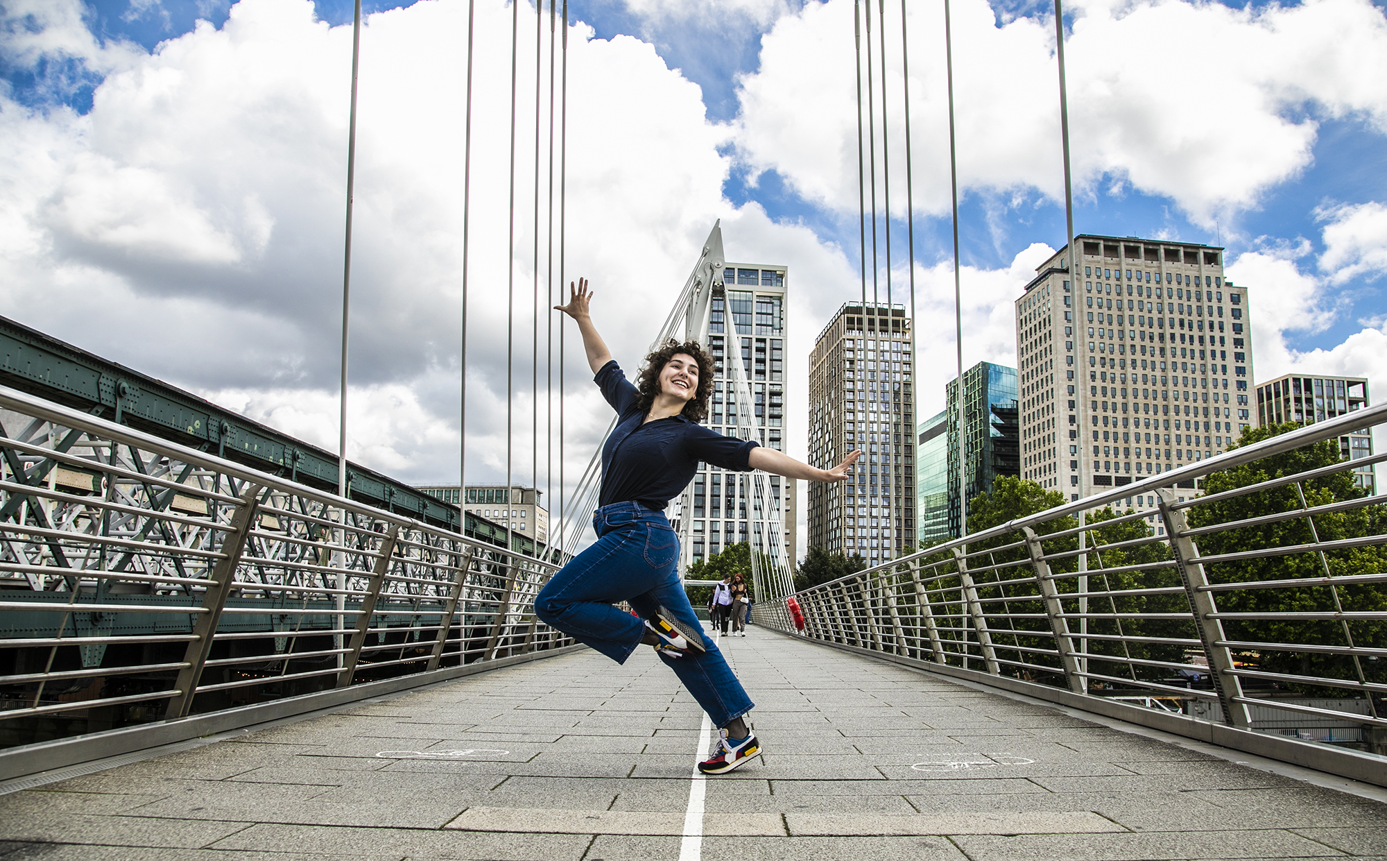 Young dancer with knee up one arm in the air one are to the side dancing on London Bridge. White female with short brown curly hair, wearing black shirt and blue jeans. Young dancer with knee up one arm in the air one are to the side dancing on London Bridge. White female with short brown curly hair, wearing black shirt and blue jeans.