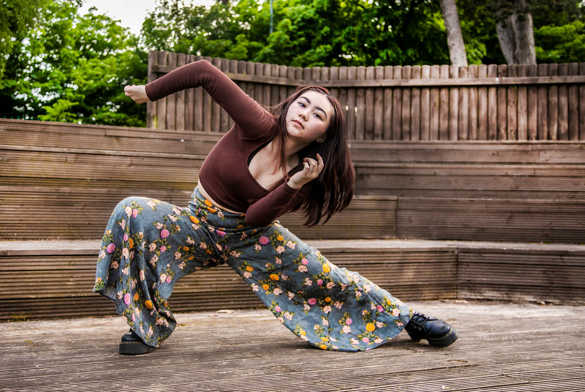 global majority female dancer lunging to the left with albow pointed in the air. In front of soft focus trees and wooden gate. Wearing brown long sleeved t-shirt and flowered flared trousers. global majority female dancer lunging to the left with albow pointed in the air. In front of soft focus trees and wooden gate. Wearing brown long sleeved t-shirt and flowered flared trousers.