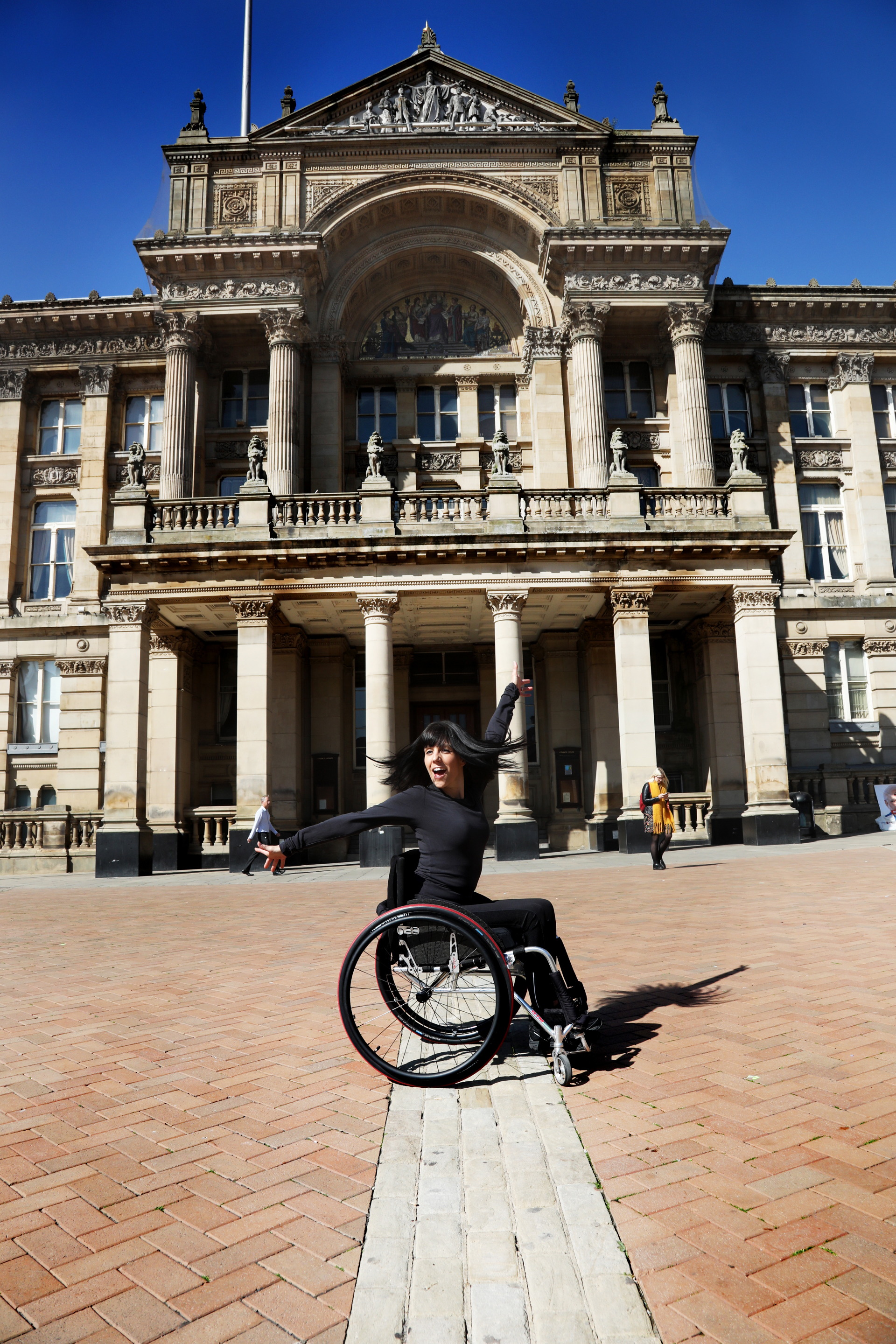 White brunette female dancer wearing all black in a wheel chair flicking her hair with one arms stretched above her head and the other out to the side. Outside a large pillared building. White brunette female dancer wearing all black in a wheel chair flicking her hair with one arms stretched above her head and the other out to the side. Outside a large pillared building.