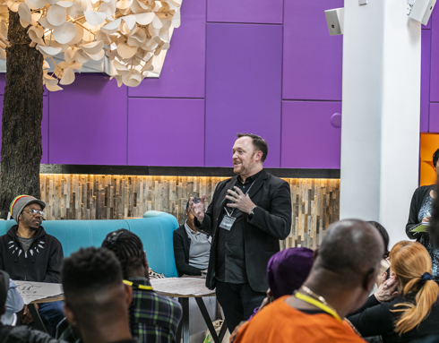 Centre of the image Andrew Hurst director of One Dance UK, white male standing in a colourful piurple room speaking to a group of people mixed genders, ages and race sitting and standing up around him. Centre of the image Andrew Hurst director of One Dance UK, white male standing in a colourful piurple room speaking to a group of people mixed genders, ages and race sitting and standing up around him.