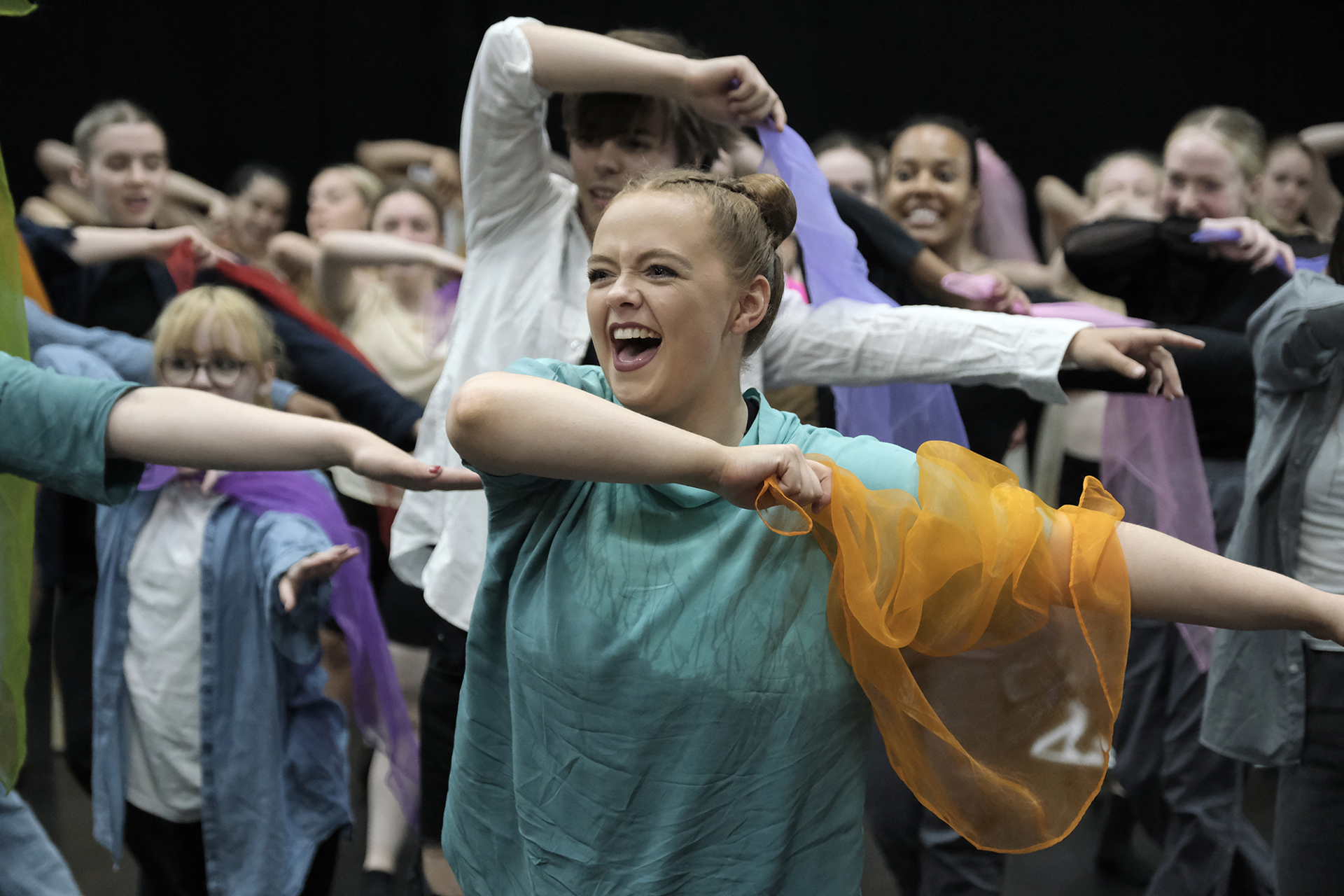 young white female dancer with red hair in a bun wearing blue top with a big happy face, holding an orange scarf with arms to the right with other young dancers behind doing the same movement. All in colourful clothes holding scarfs. young white female dancer with red hair in a bun wearing blue top with a big happy face, holding an orange scarf with arms to the right with other young dancers behind doing the same movement. All in colourful clothes holding scarfs.