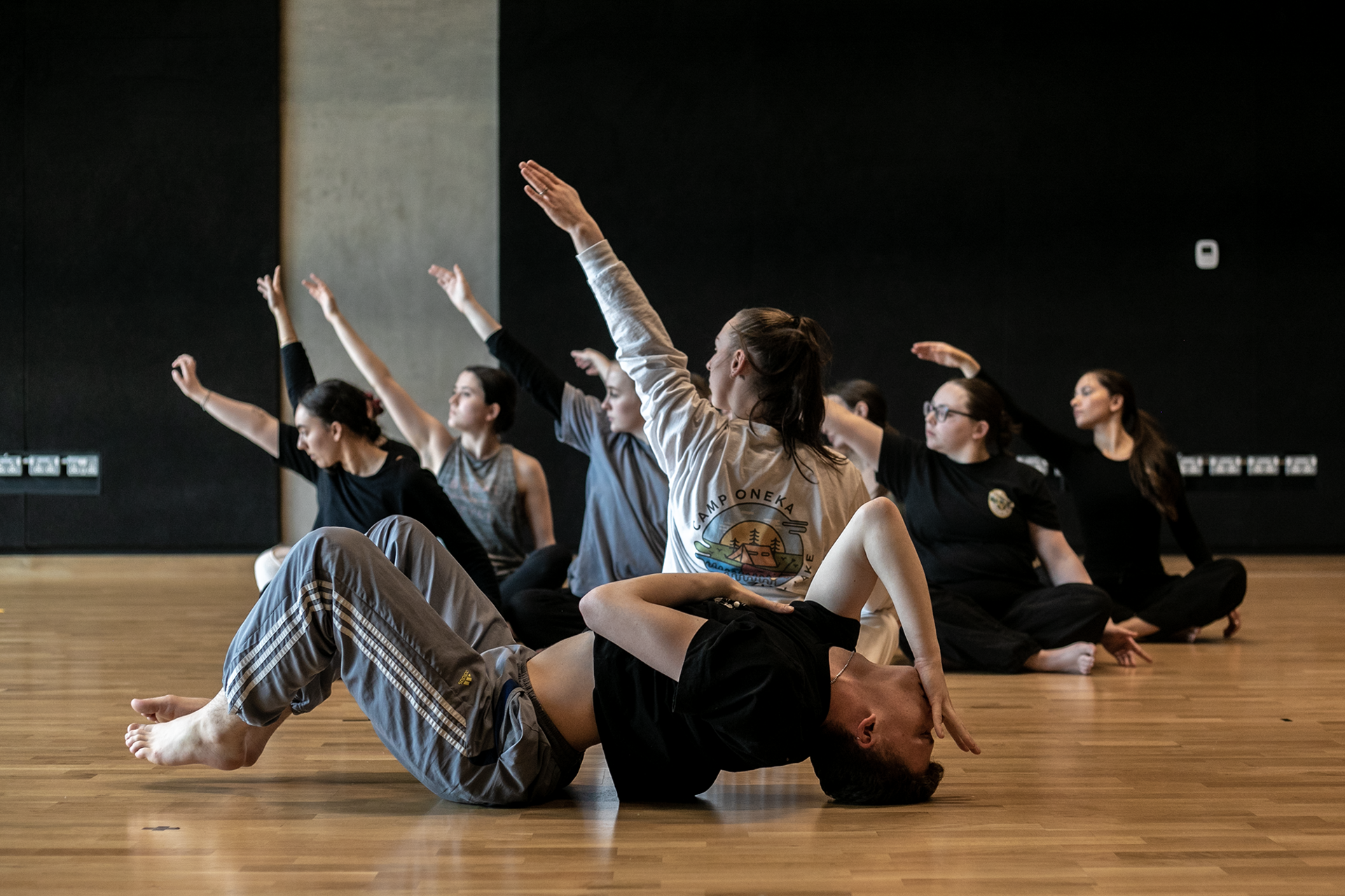 7 female dancers sitting crossed legged with one arm in the air. one male dancer infront laying on the floor with knees up arched back and hand infront of face. In a dance stucio with wooden floors and black walls 7 female dancers sitting crossed legged with one arm in the air. one male dancer infront laying on the floor with knees up arched back and hand infront of face. In a dance stucio with wooden floors and black walls