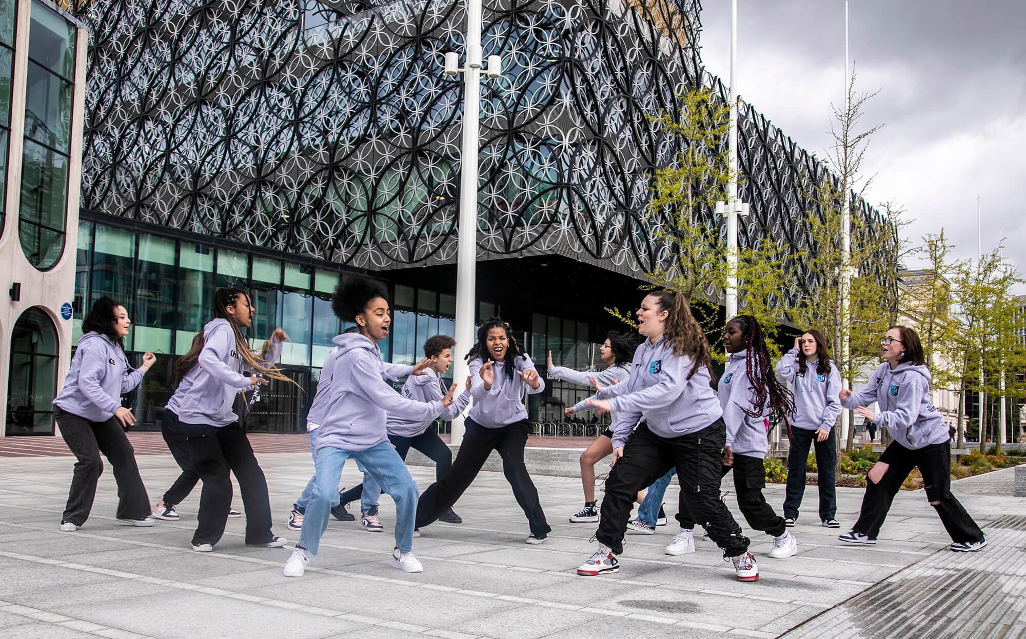 Group of teenage female dancers performing street dance infront of large building with circles. All wearing matching grey hoodies Group of teenage female dancers performing street dance infront of large building with circles. All wearing matching grey hoodies