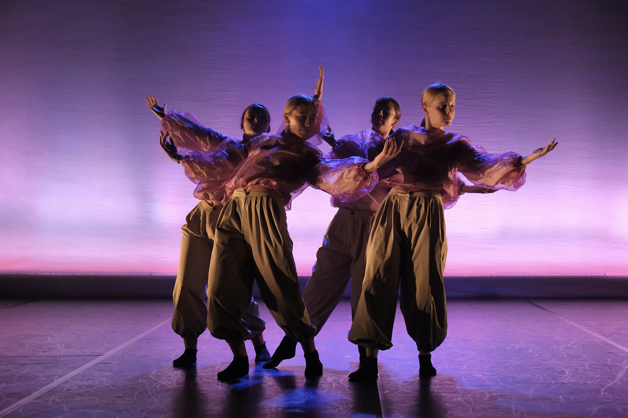 4 female dancers wearing baggy pink tops and cargo trousers all huddled together with arms stretched out leaning towards eachother. Stage lighting is pink and purple 4 female dancers wearing baggy pink tops and cargo trousers all huddled together with arms stretched out leaning towards eachother. Stage lighting is pink and purple