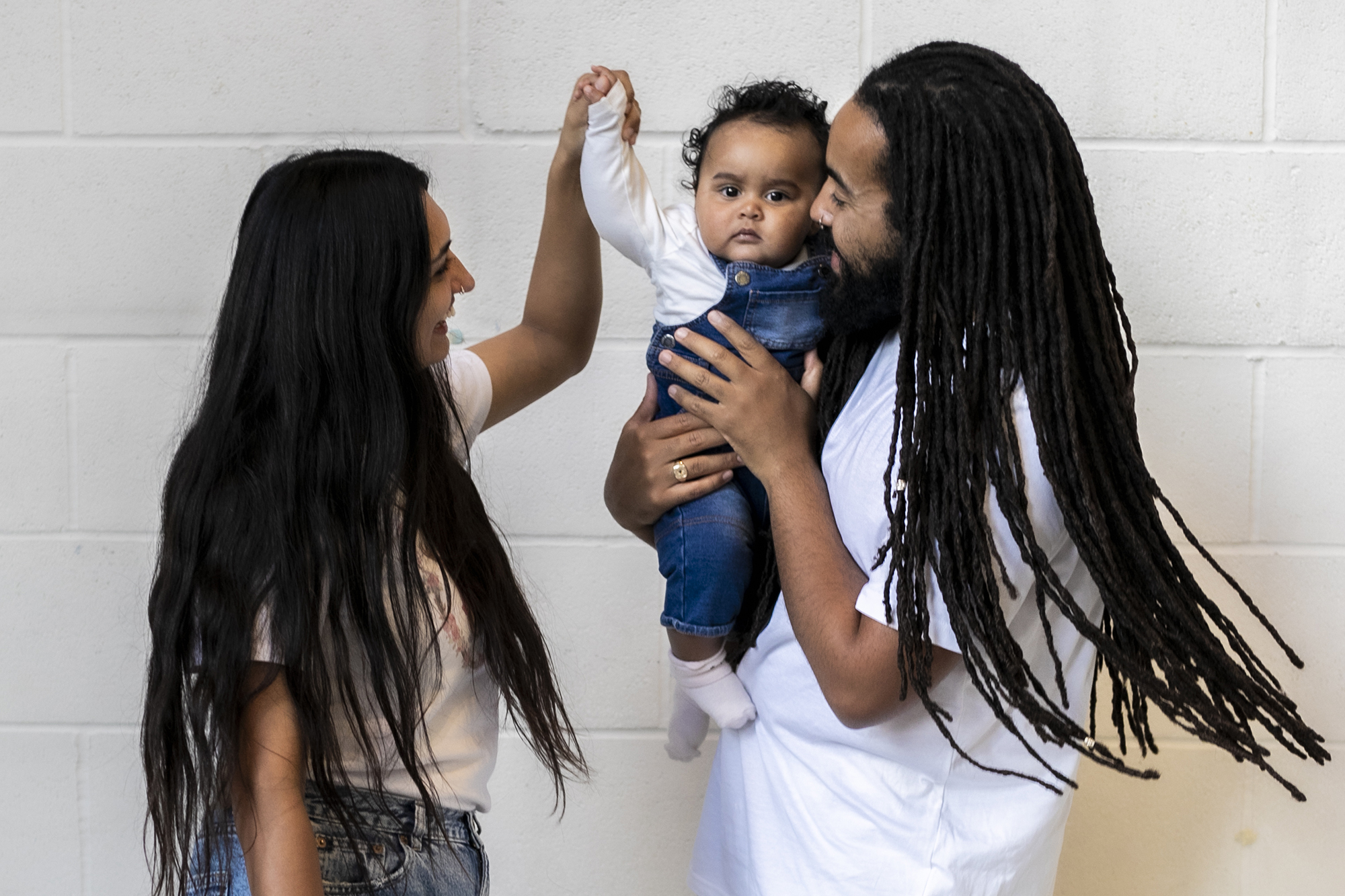 Family of 3. Global majority male with long dreadlocked hair holding baby, global majority female with long black hair, holding the babies hand as they dance. Wearing blue jeans, trainers and white tshirts infront of white brick wall. 