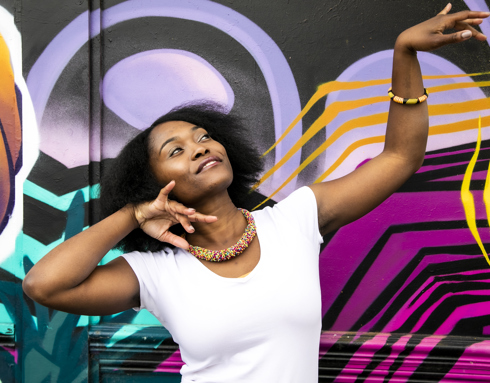 Global majority female dancer with afro, holding one arm out to the top right of the image with eyes looking at a limp wrist and the other hand next to her face. Wearing white top and colourful beaded necklace in fornt of coloured graffiti wall Global majority female dancer with afro, holding one arm out to the top right of the image with eyes looking at a limp wrist and the other hand next to her face. Wearing white top and colourful beaded necklace in fornt of coloured graffiti wall