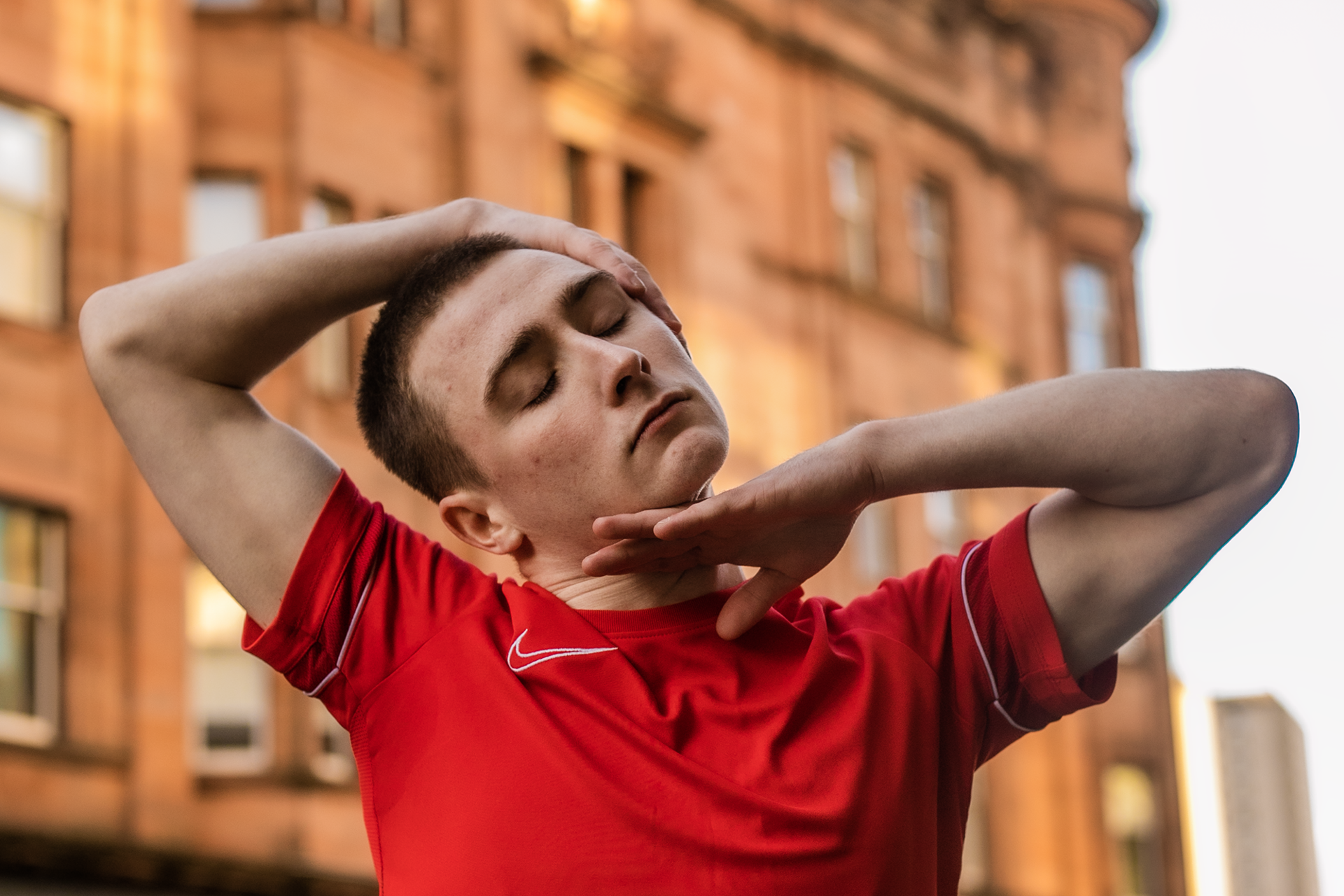 white male dancer with shaved head and red t shirt. On the street infront of orange building with hands around the face 