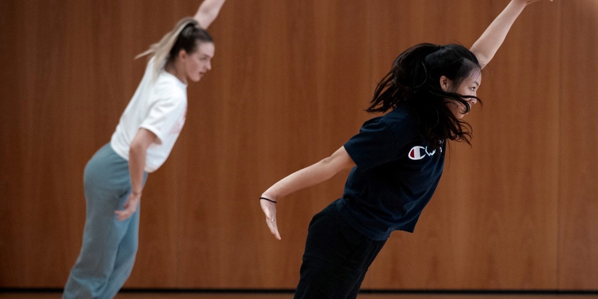 two young female dancers leaning diagonally with one arm up and other by their side. In front of wooden wall