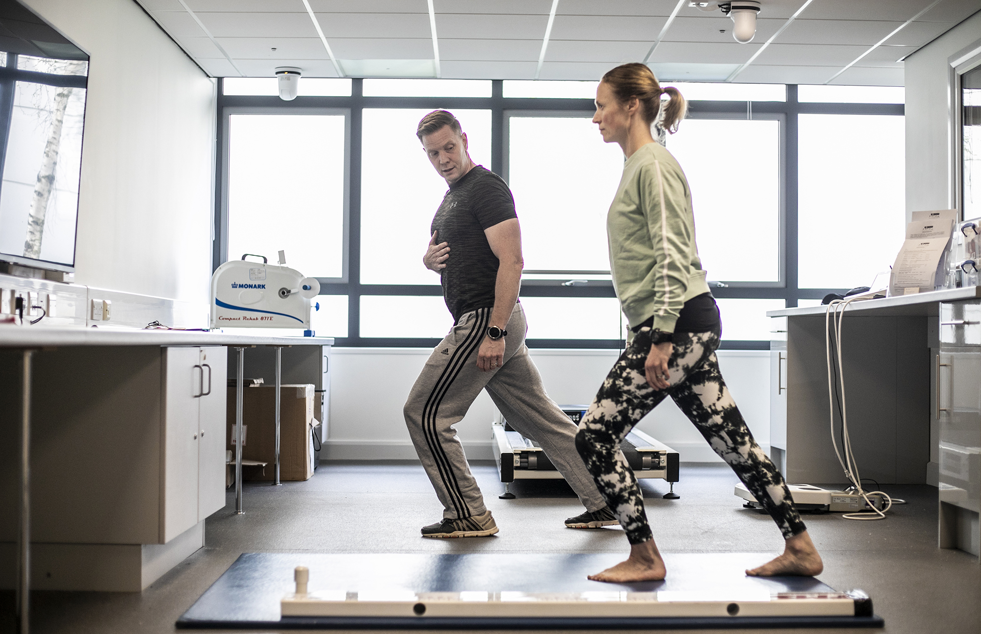 white male physio lunging forward holding his stomach, with white female dancer mirroring his movement. Both wearing excersise clothes in a bright healthcare labratory with large windows in the background. white male physio lunging forward holding his stomach, with white female dancer mirroring his movement. Both wearing excersise clothes in a bright healthcare labratory with large windows in the background.