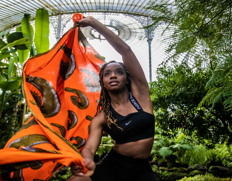 Black female dancer with long brown braids moving a bright orange scarf in the air, looking above the camera, in greenhouse surrounded by plants. Wearing black crop top and black cycling shorts. Black female dancer with long brown braids moving a bright orange scarf in the air, looking above the camera, in greenhouse surrounded by plants. Wearing black crop top and black cycling shorts.