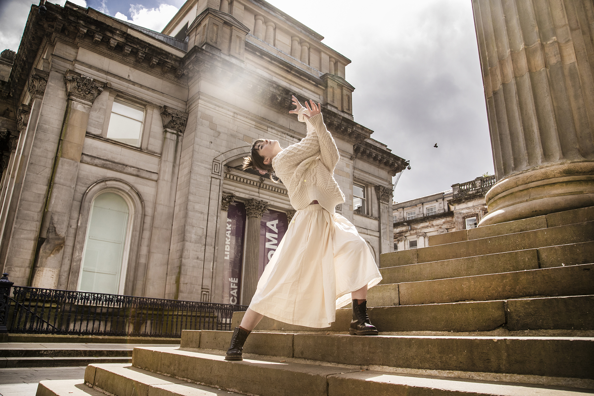 dancer lunging on steps of Glasgow council building with large pillars. White female dancer with brown hair arms interlinked towards the sky. White cable knit jumper and white midi skirt. dancer lunging on steps of Glasgow council building with large pillars. White female dancer with brown hair arms interlinked towards the sky. White cable knit jumper and white midi skirt.