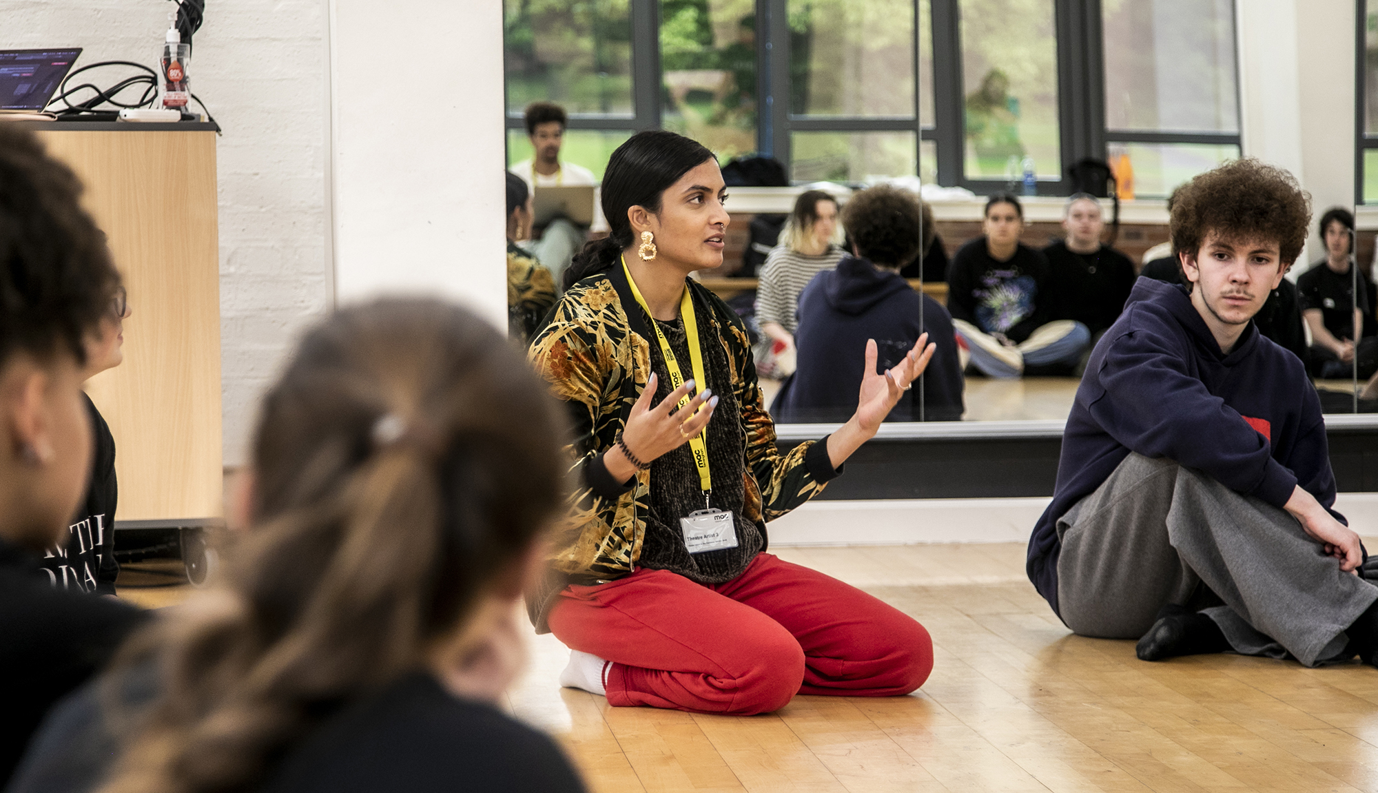golbal majority dance teacher sitting on her knees speaking to a room full of dance students gesturing her hands, with mirror reflecting the students behind her. Wearing red trousers and colourful jacket. golbal majority dance teacher sitting on her knees speaking to a room full of dance students gesturing her hands, with mirror reflecting the students behind her. Wearing red trousers and colourful jacket.