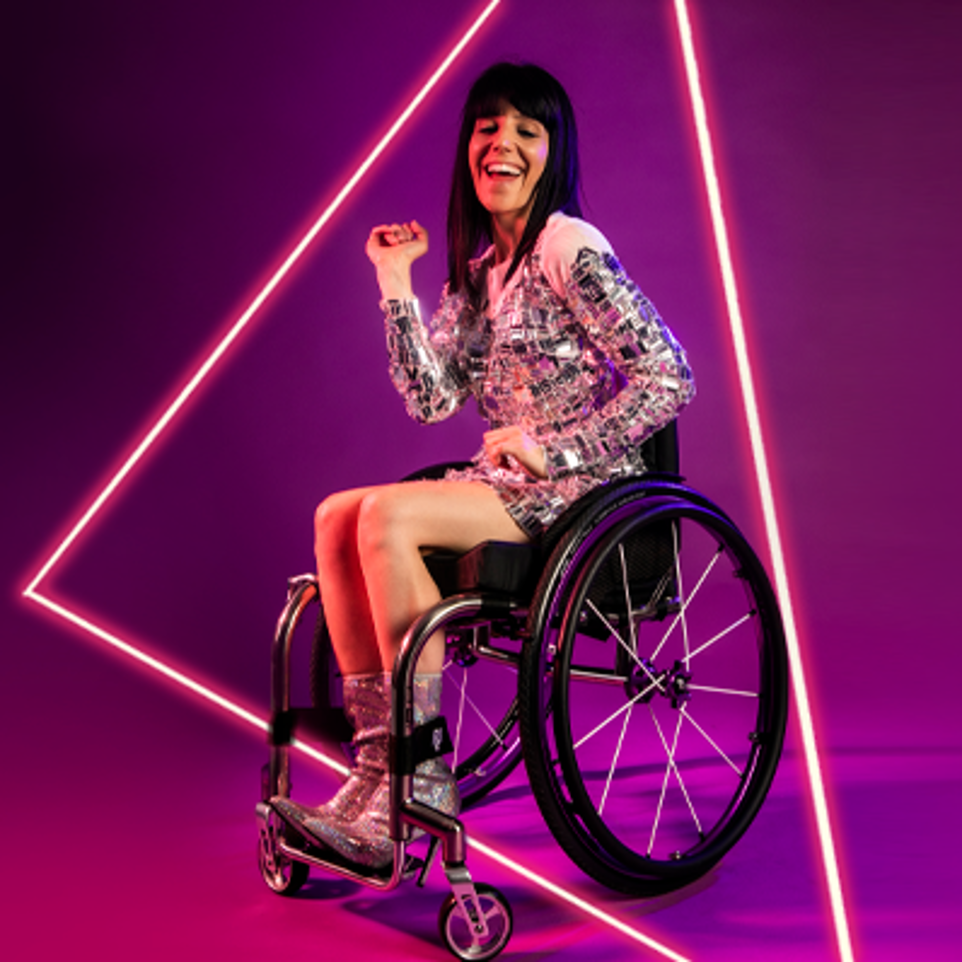 White female wheelchair dancer with black hair. With one arm up and one down with wheel chair side on. Wearing silver sparkle dress and silver high heeled shoes in front of hot pink studio background. White female wheelchair dancer with black hair. With one arm up and one down with wheel chair side on. Wearing silver sparkle dress and silver high heeled shoes in front of hot pink studio background.