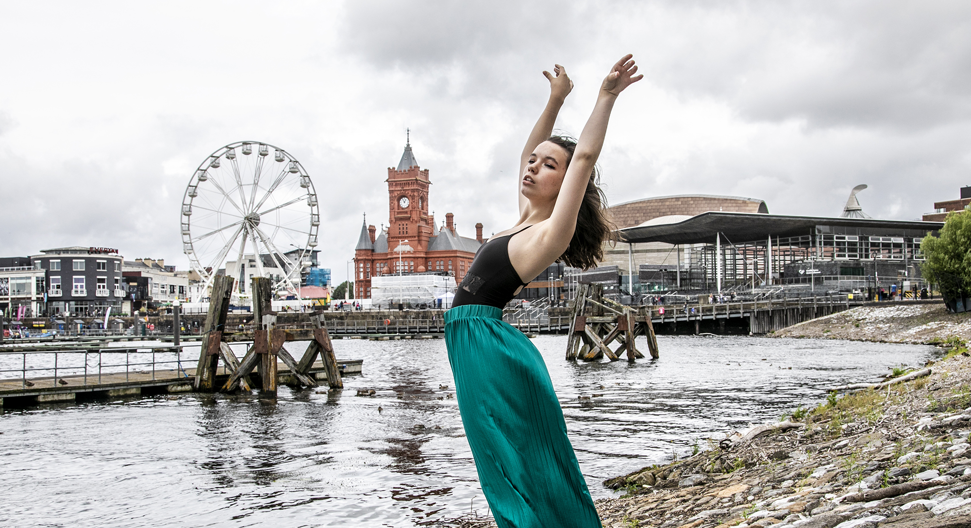 dancer with arched back and arms above head infront of Cardiff bay skyline. Young white female wearing black leotard and green silk trousers dancer with arched back and arms above head infront of Cardiff bay skyline. Young white female wearing black leotard and green silk trousers